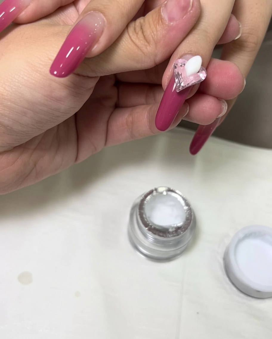 A bead of white 3D sculpting gel being placed on the nail next to a pink heart crystal.