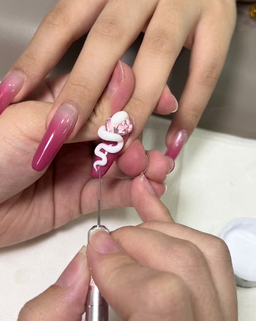 A close-up of a hand using a metal precision tool to shape a white 3D gel snake over a pink gemstone on a magenta ombre nail.
