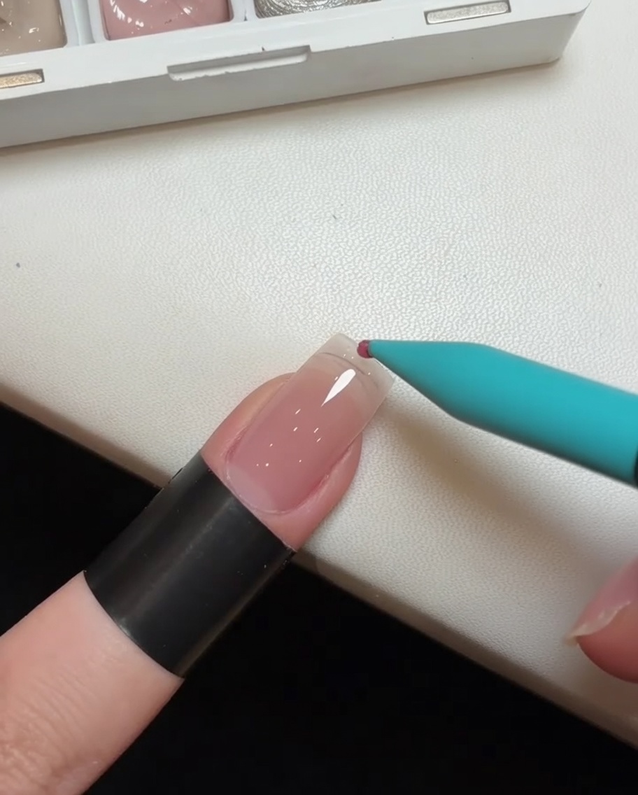 A close-up view of a detail brush applying a dot of pink gel polish to the tip of a clear nail extension.