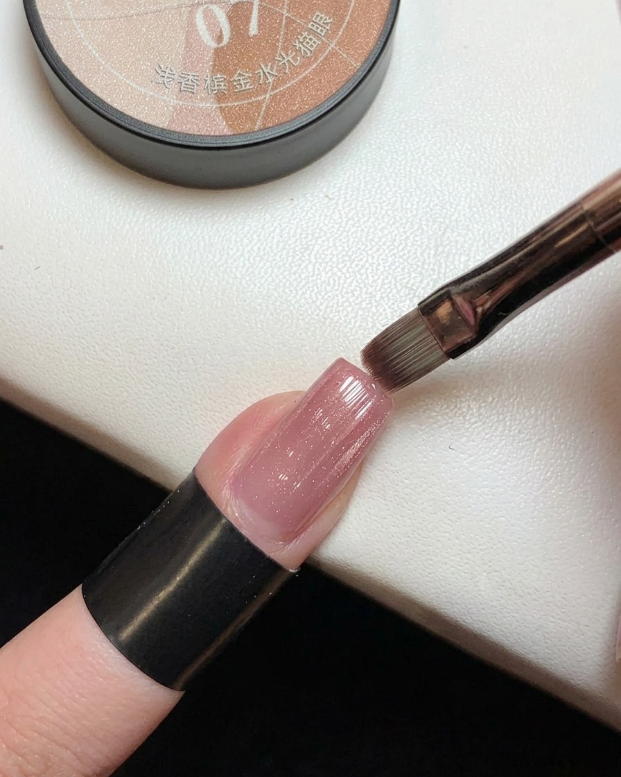 A brush applying sheer pink magnetic gel with gold shimmer over a previously cured pink gradient nail.