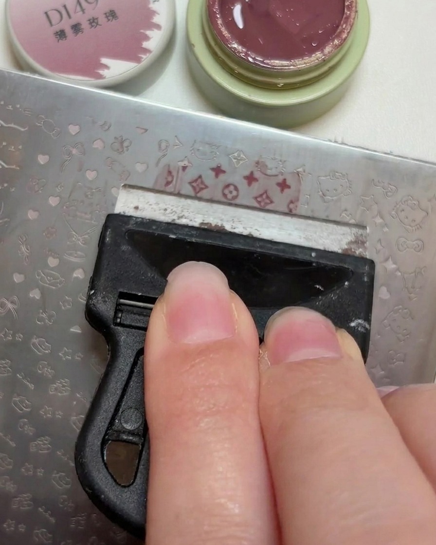 A plastic scraper leveling a small dollop of dusty rose gel polish across the engraved patterns on a metal nail stamping plate.