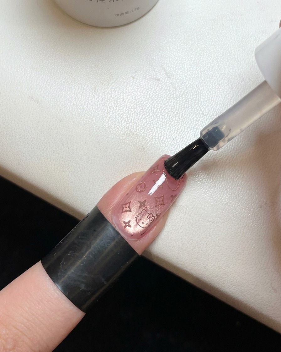 A brush applying a clear, glossy top coat over a nail that has a pink Hello Kitty pattern.