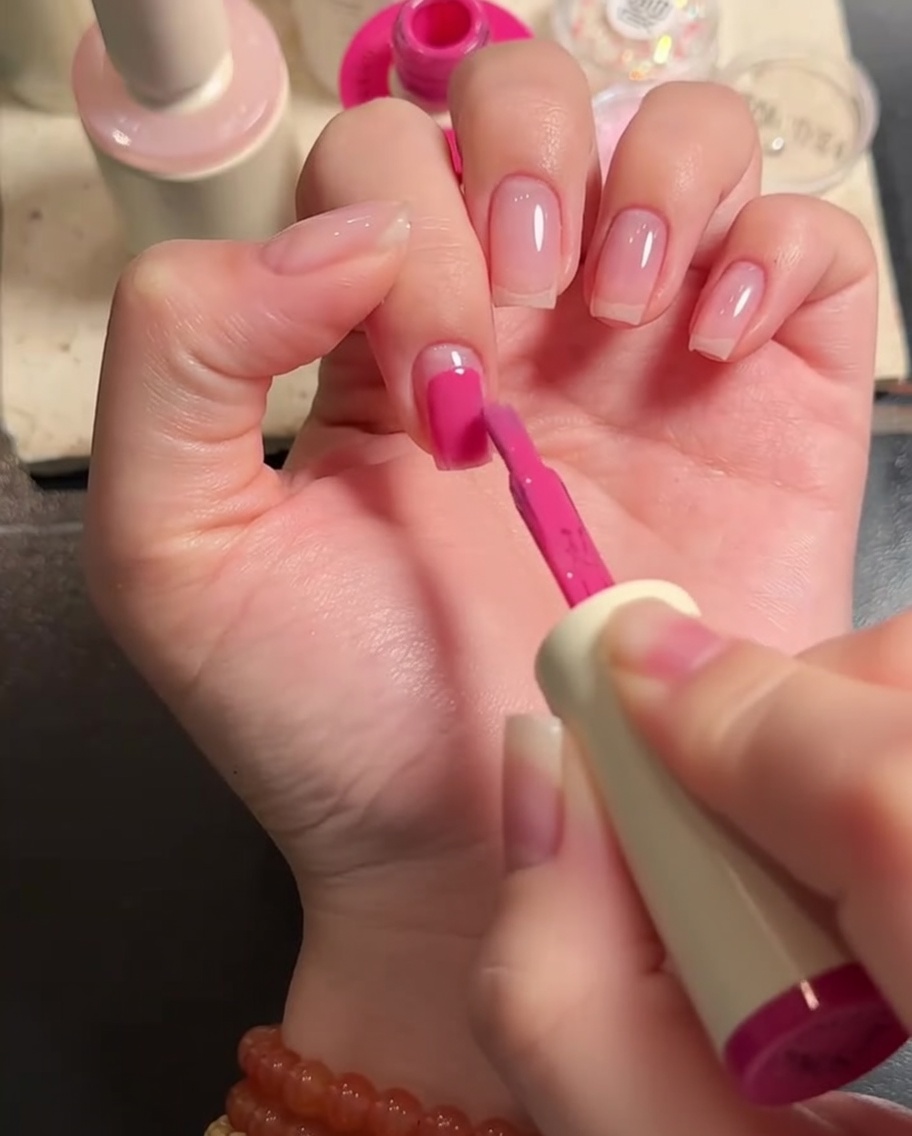 A close-up of a flat nail brush applying pink gel polish to the surface of a fingernail.