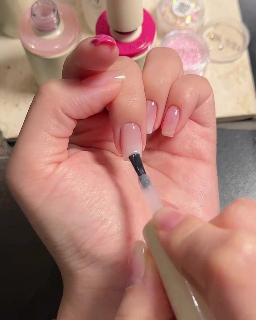 A brush applying a coat of sheer nude gel polish over the middle and ring fingers to blend the gradient.
