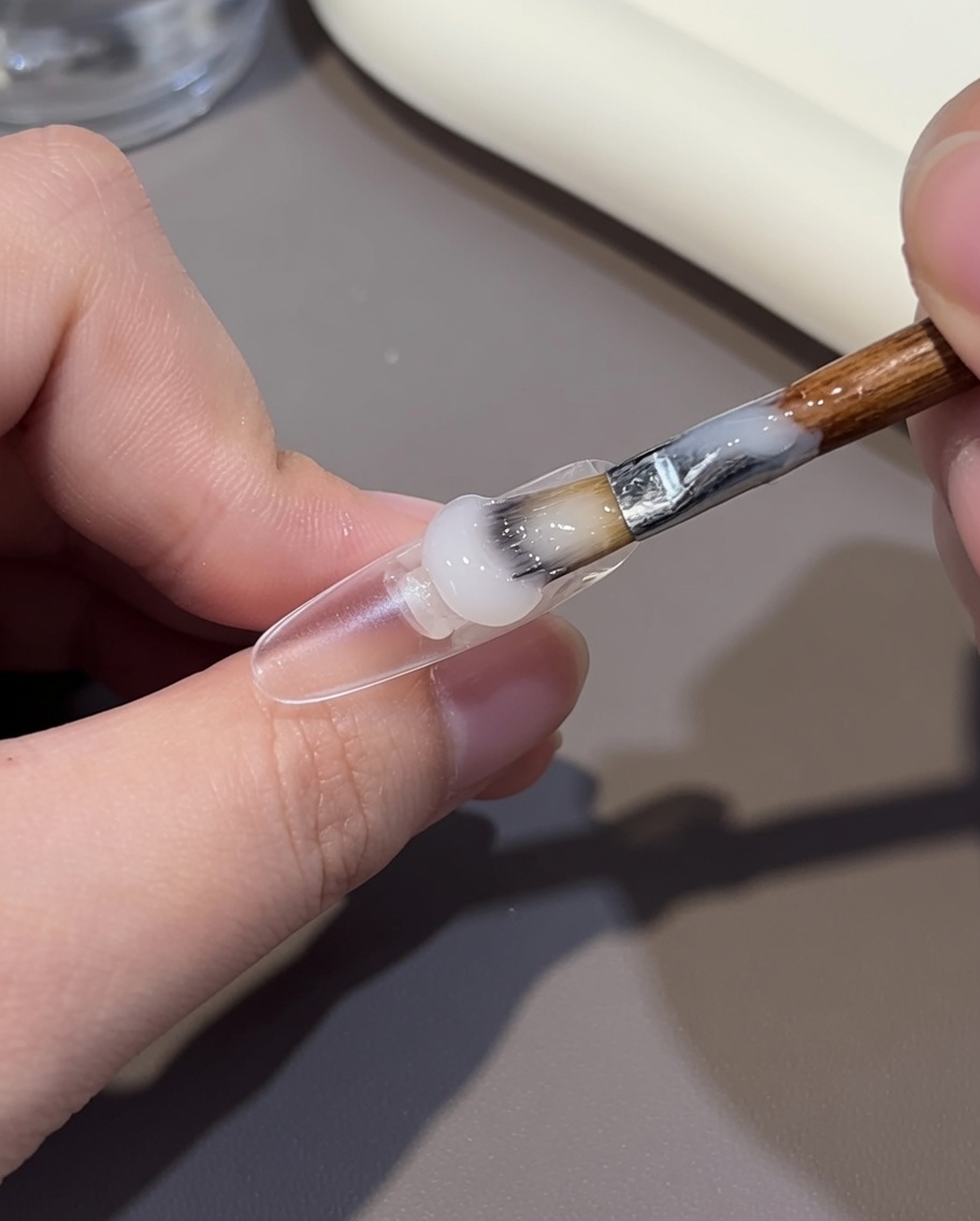 A close-up of a brush applying a nude-colored gel to the cuticle end of a clear nail tip.