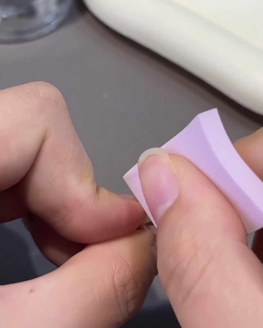 A close-up view of a pink ombre nail being coated with a second layer of pink gel polish using a small brush.