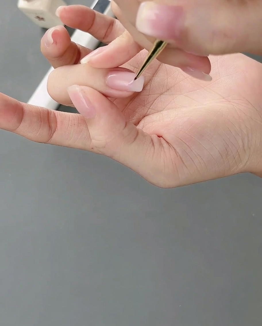 A fine-tipped nail art brush applying light pink gel onto a nail to form irregular color blocks.