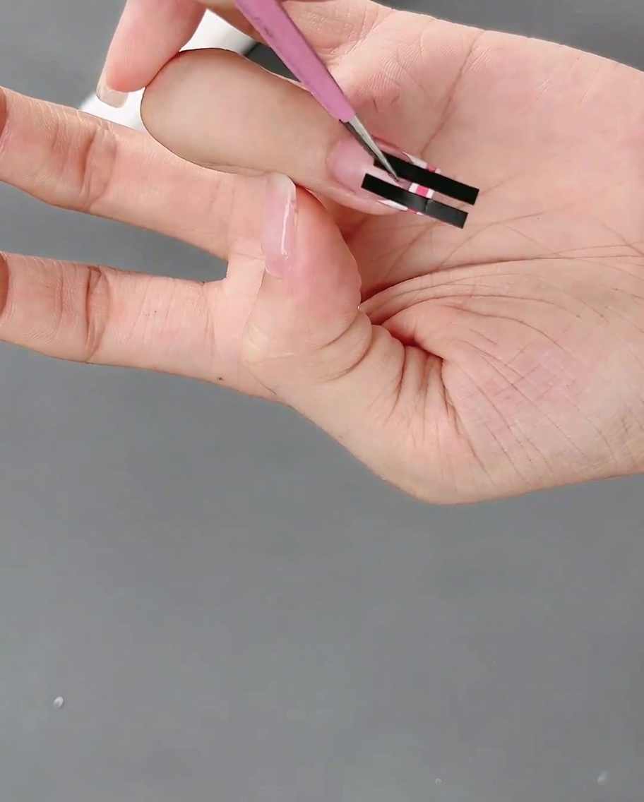 Tweezers gently peeling away black striping tape from a nail to reveal a pink and white checkered grid pattern.