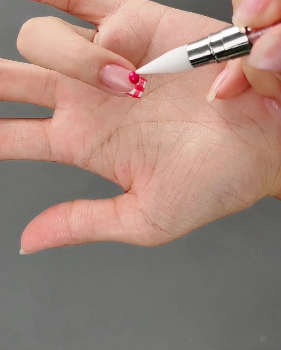 A silicone tool pointing at a small, round bead of dark pink gel resting on a checkered nail tip.
