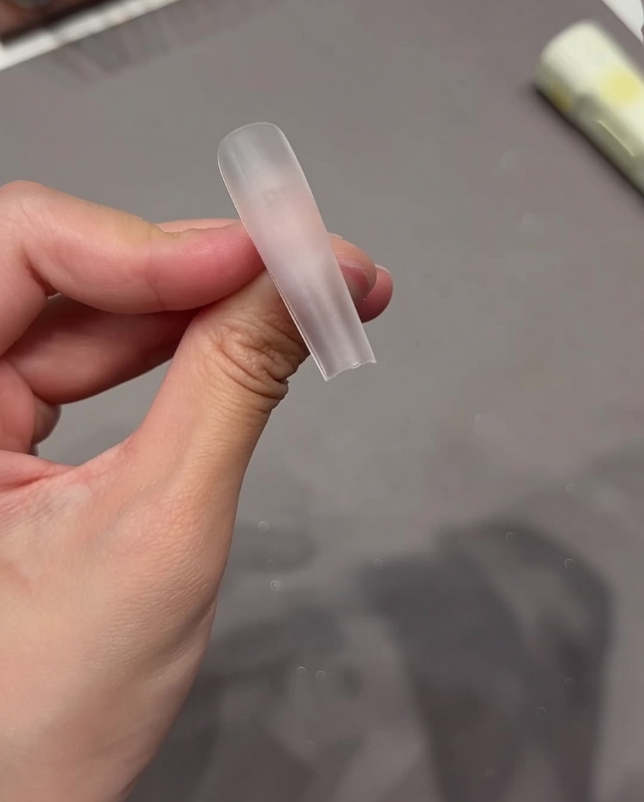 A close-up of a hand applying a transparent sticky tab to the surface of a clear nail tip.