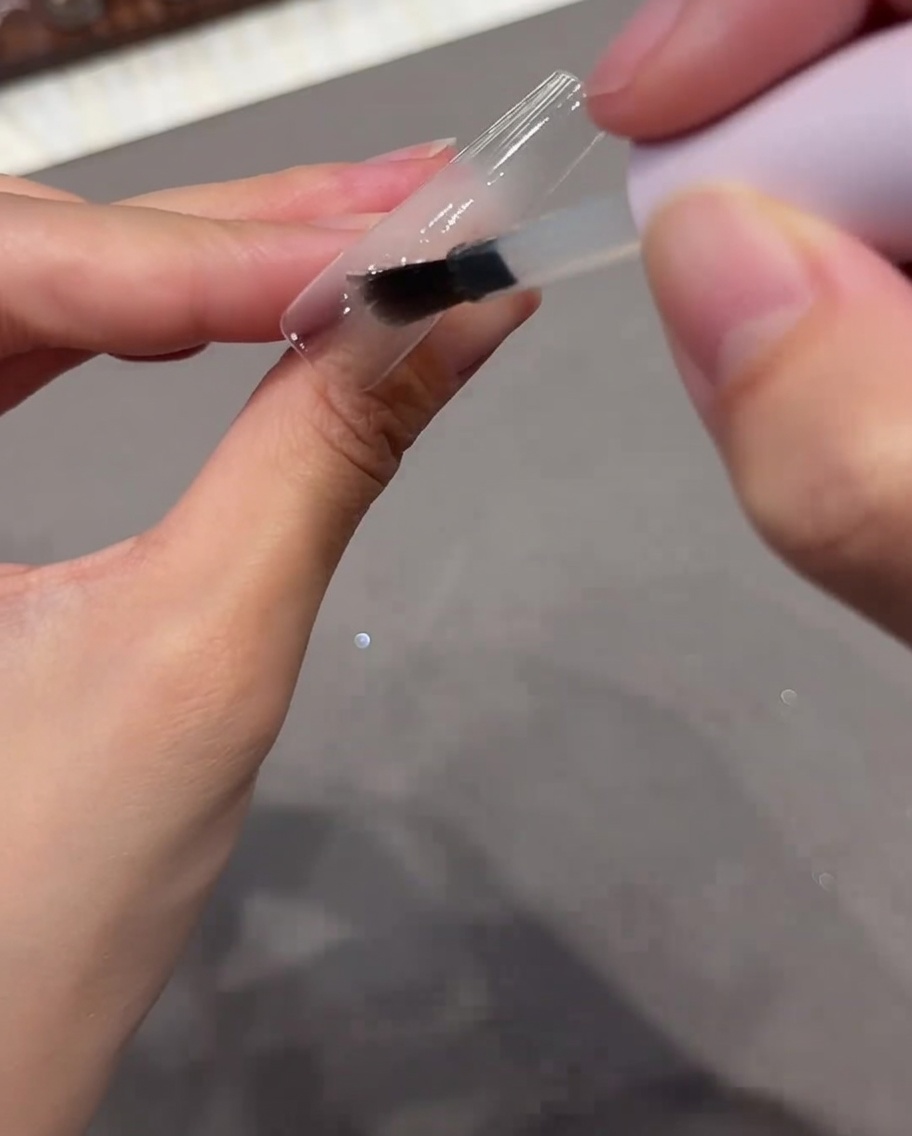 A brush applying a clear base coat gel onto a clear nail tip.
