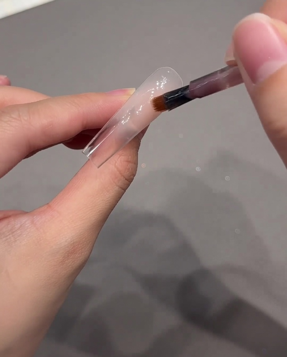 A nail art brush applying a dot of nude pink gel to the cuticle area of the nail tip.
