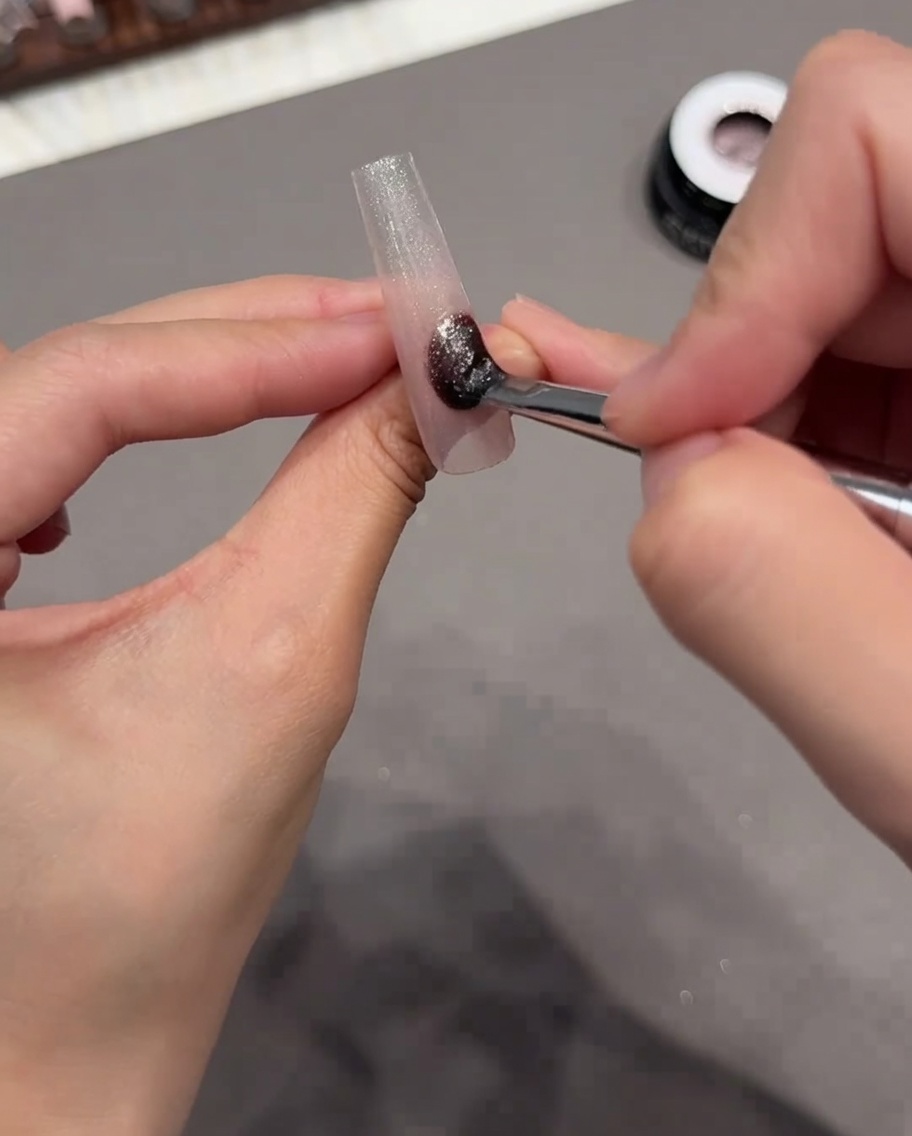 A brush applying a shimmering cat eye gel polish over the entire surface of the nail tip.
