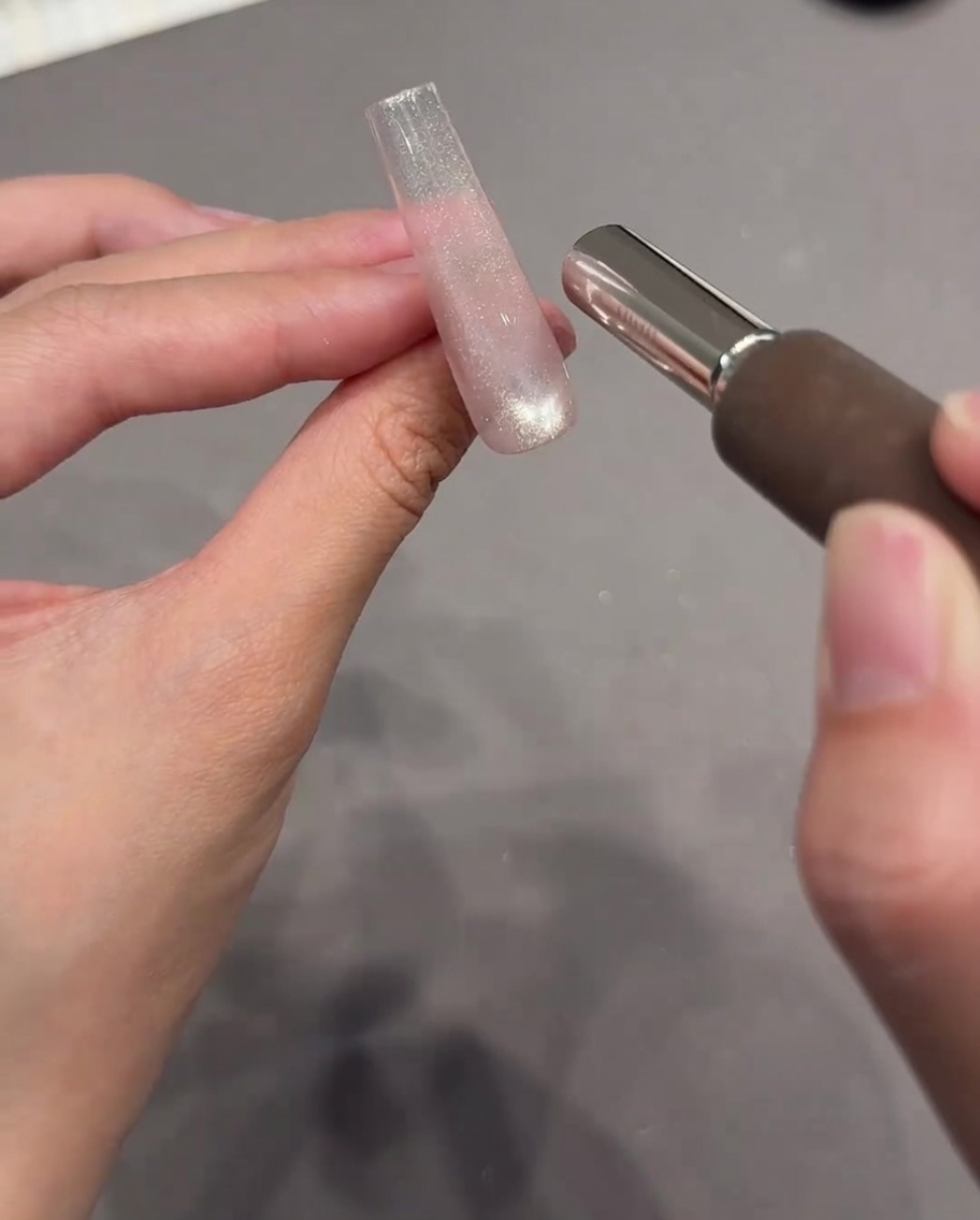 A hand holding a cylindrical magnet tool close to a clear nail extension coated with wet, sparkly cat eye gel.