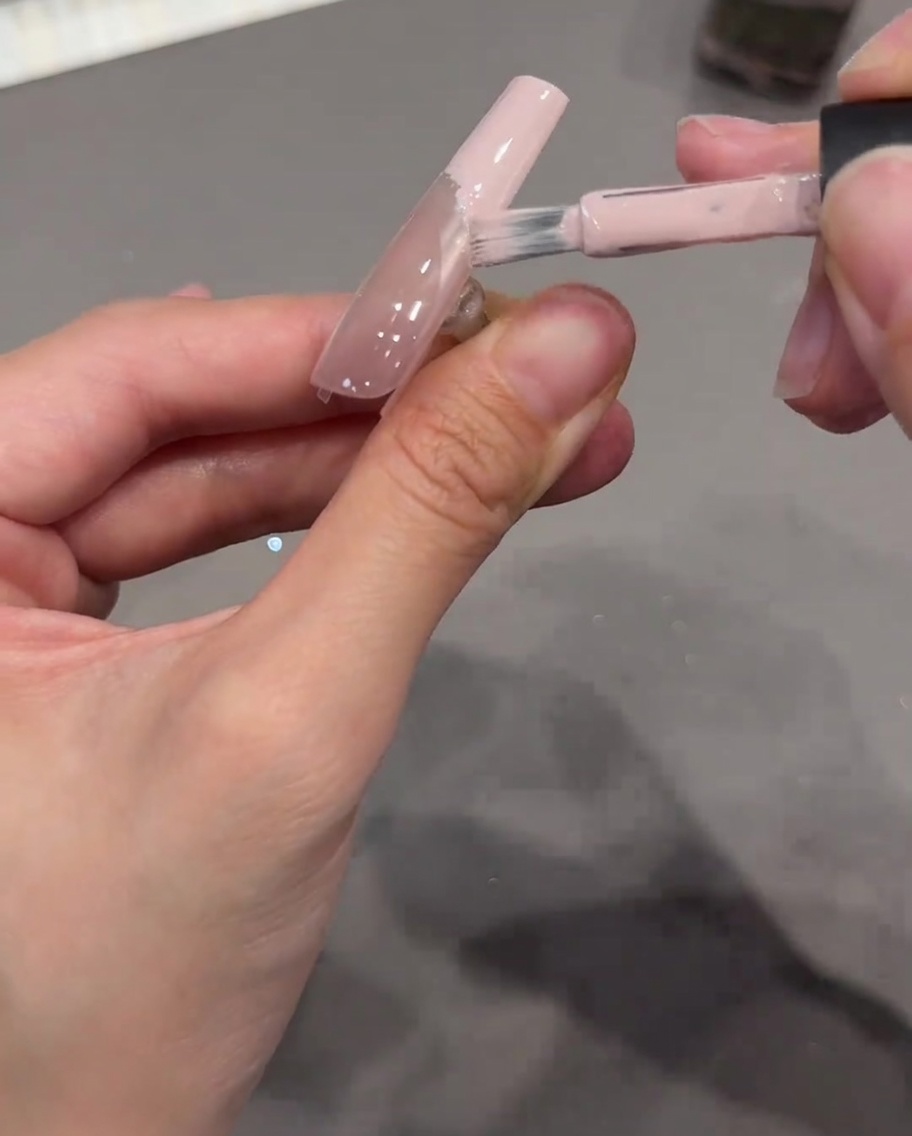 A nail brush applying opaque nude pink gel onto the tip of a nail, using a clear curved sticker as a clean boundary.