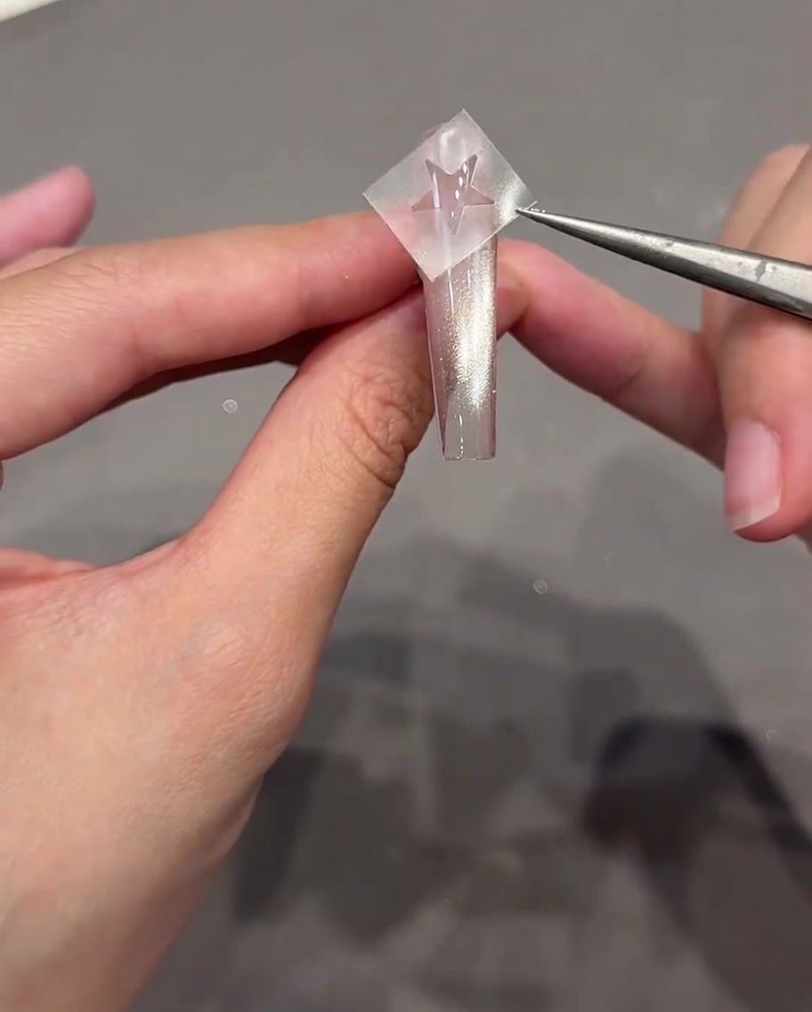 A pair of tweezers holding a star-shaped sticker just above a clear, cat-eye style nail.