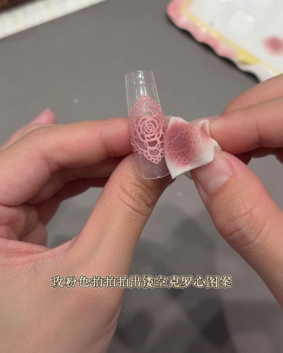 A close-up view of a hand using a small makeup sponge to dab over a lace-like sticker on a clear nail tip.