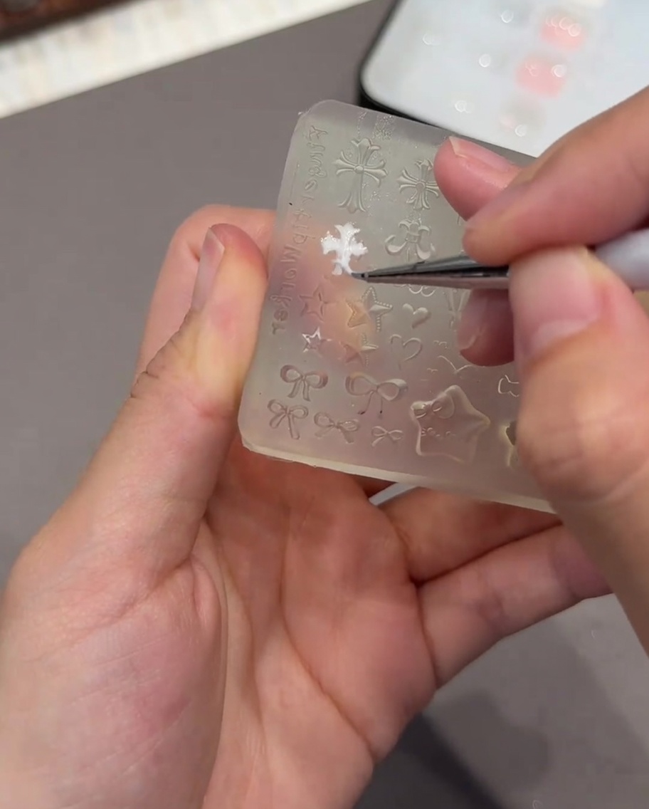 A pair of tweezers holding a small white 3D heart-shaped mold, preparing to place it onto a nail.