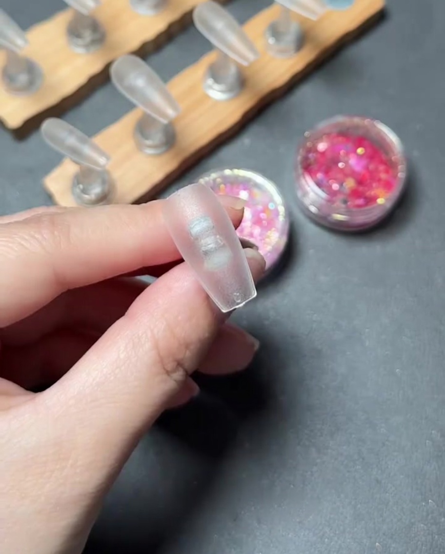A close-up of a brush applying clear base gel onto the surface of a clear plastic nail tip.