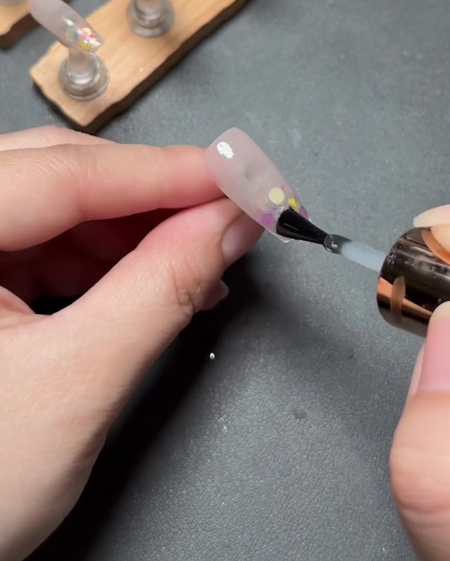 A thin, clear layer of gel being brushed over the finished pink gradient and glitter nail.
