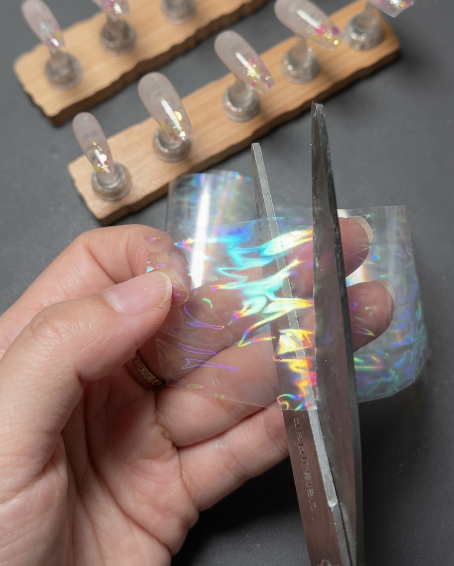 Hands using scissors to cut a small rectangular piece from a strip of holographic laser aurora transfer foil.