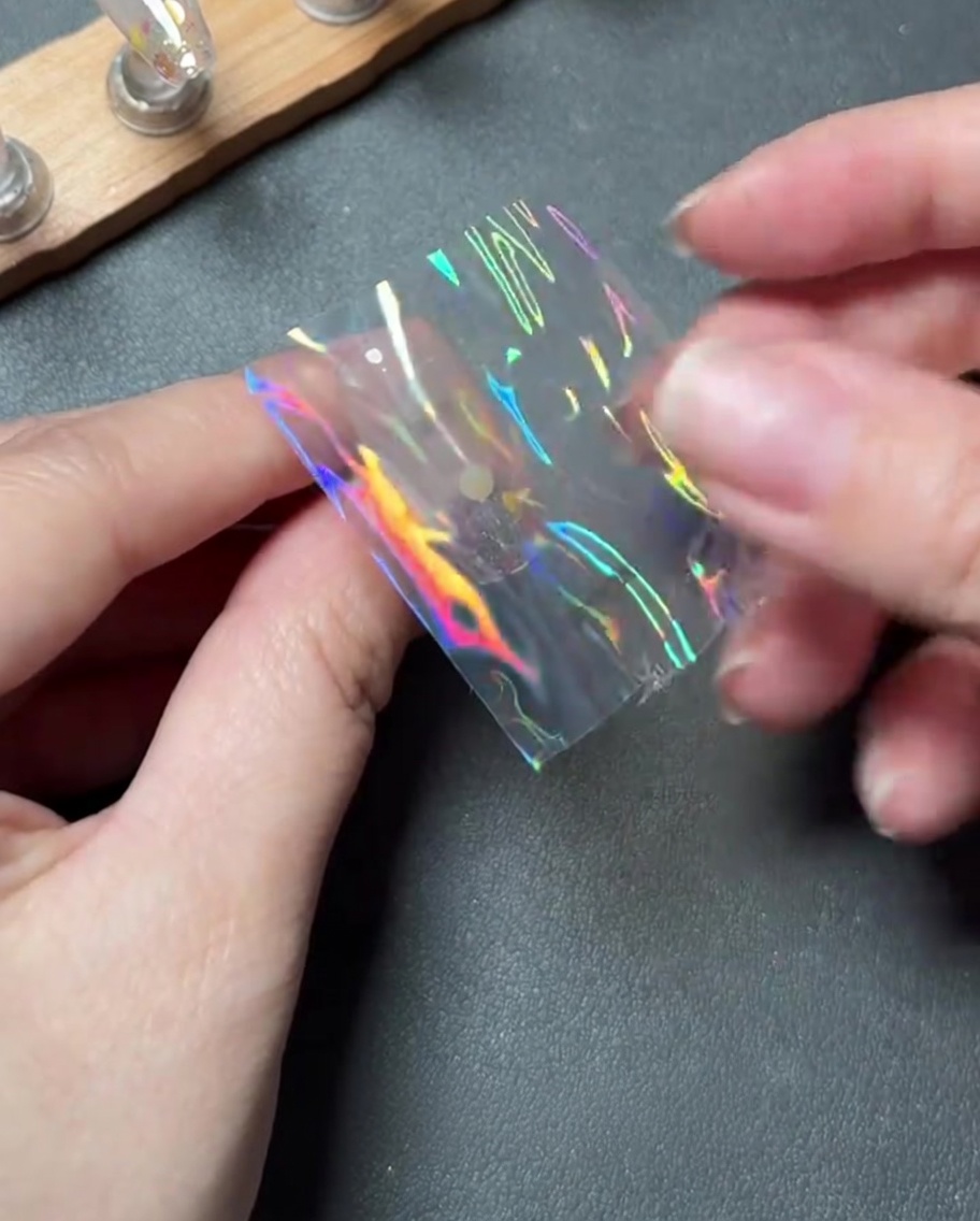 Fingers carefully placing a small piece of holographic transfer foil onto the center of a nail.