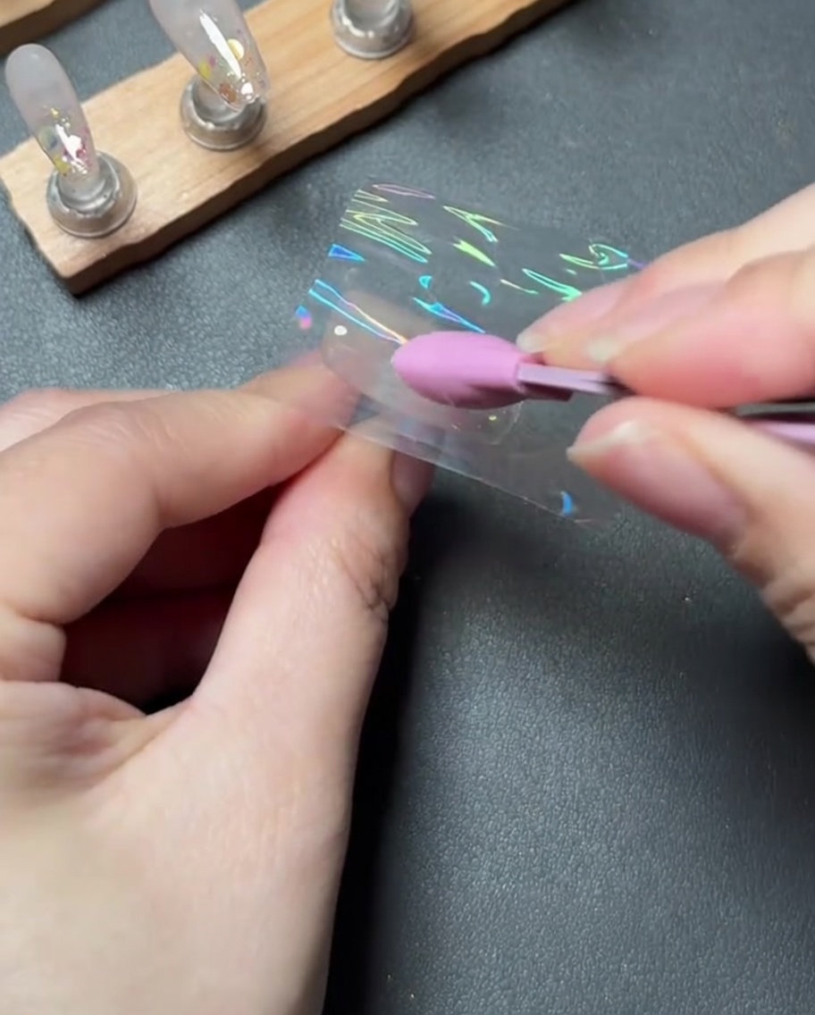 A pink silicone tool firmly pressing and smoothing a piece of holographic transfer foil onto a nail.