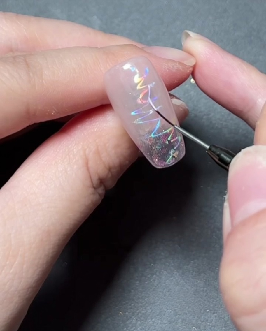 A fine liner brush painting a delicate star shape with colorful gel onto a holographic foiled nail.