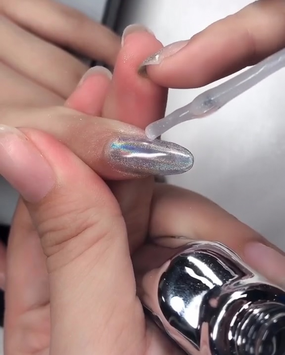 A nail brush applying a clear, viscous reinforcing gel over a chrome-finished nail, carefully coating the free edge.