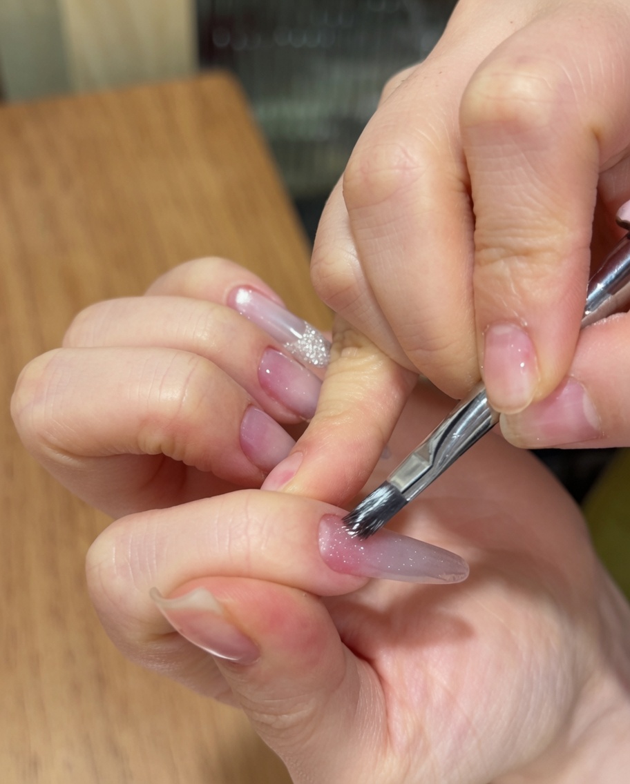 A hand using a nail brush to apply a sparkly cat eye gel polish over a long almond nail with a pink gradient base.