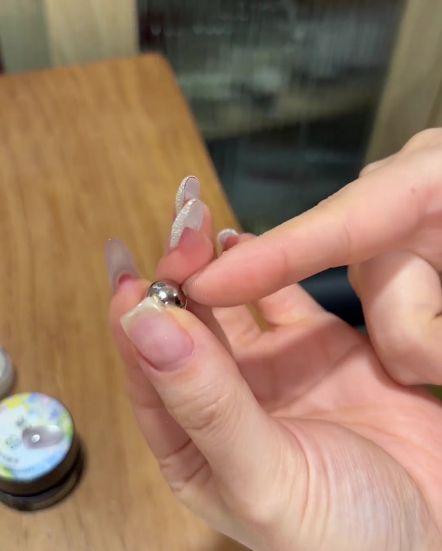 A hand holding a small spherical silver magnet closely over a fingernail painted with uncured sparkly cat eye gel.
