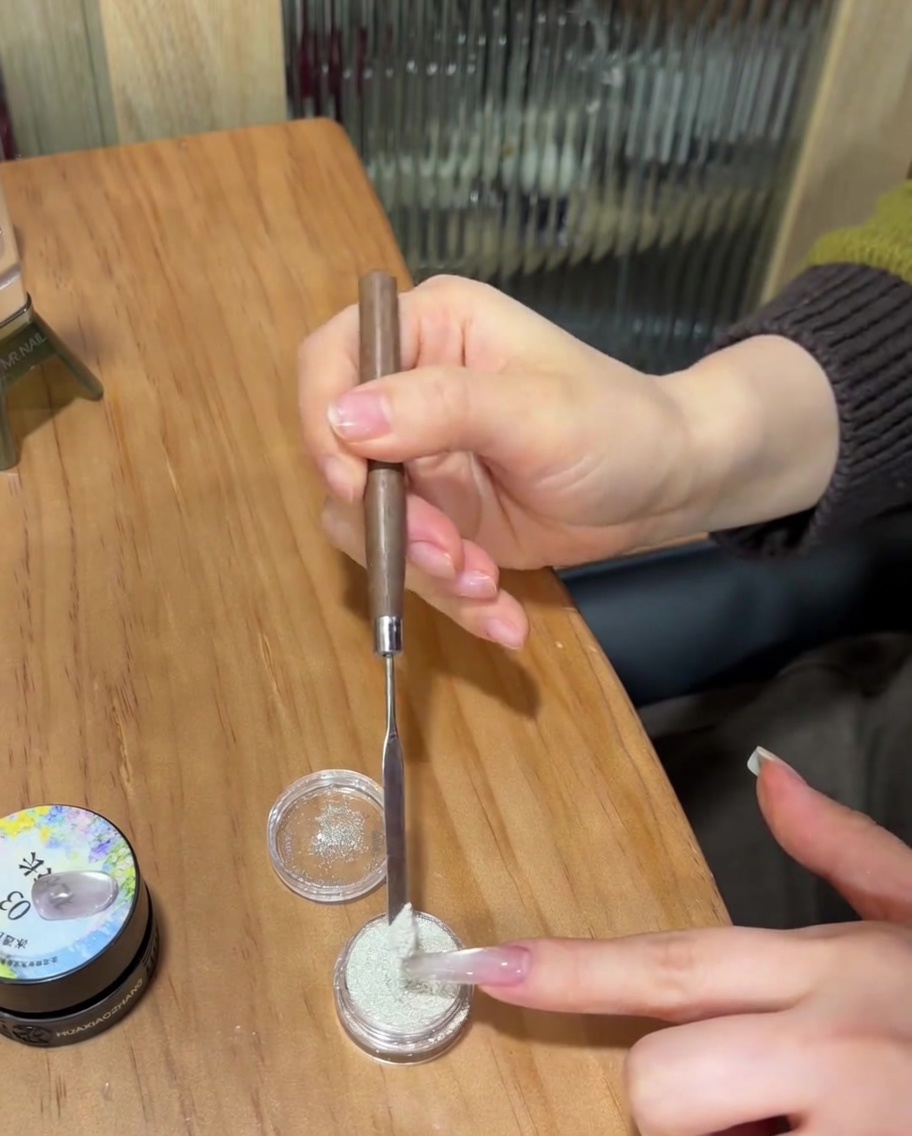 A close-up of a nail with uncured clear gel being coated in fine white sugar powder using a metal spatula.