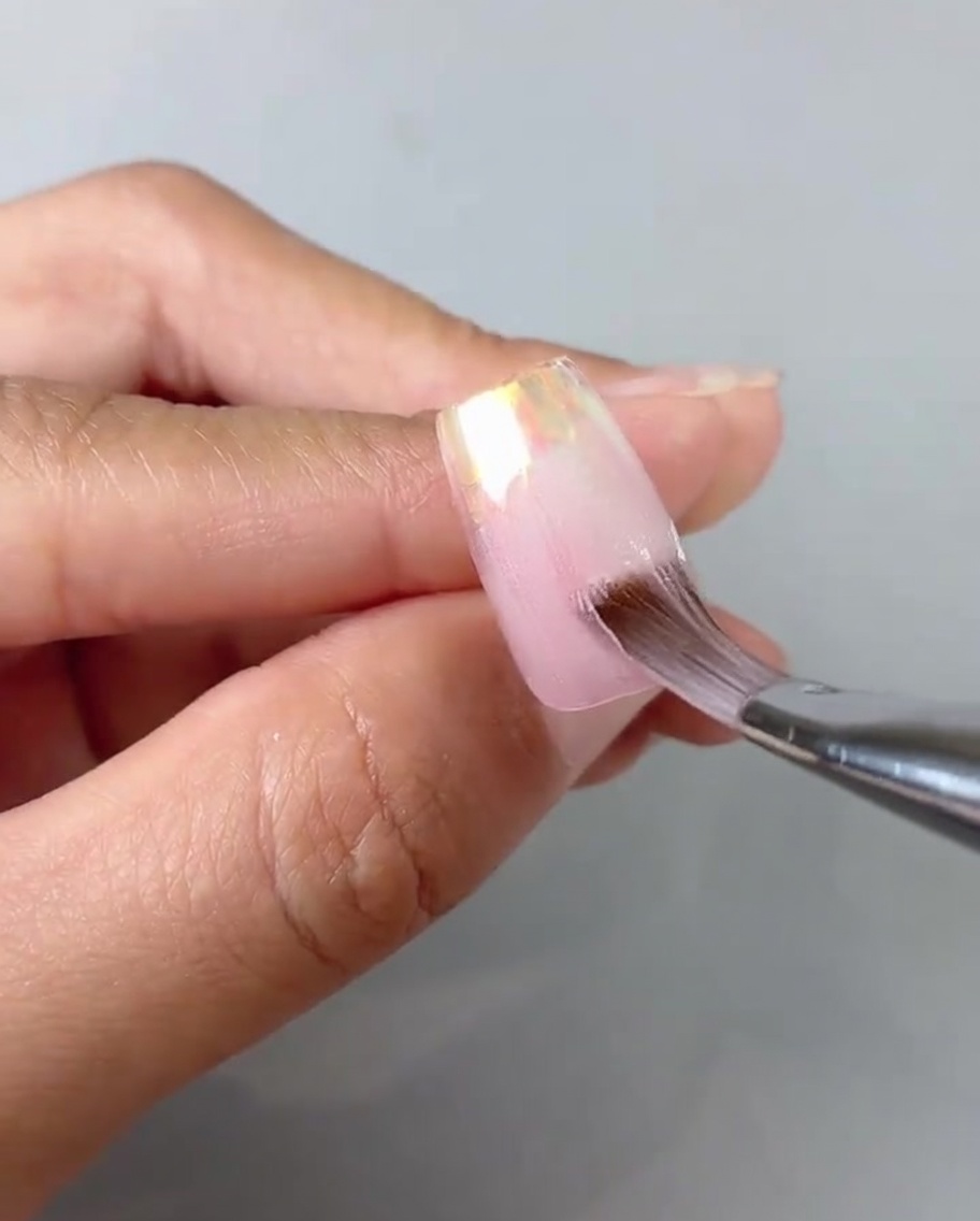 A close-up of a brush smoothing and blending sheer pink gel on a nail tip.