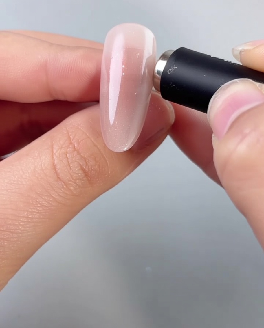 A hand holding a cylindrical magnet close to an almond-shaped nail, activating the shimmering particles in the wet cat-eye gel.