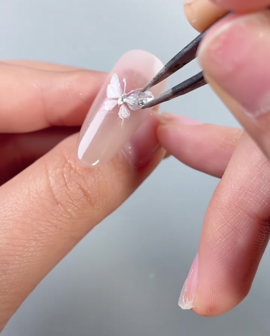 Tweezers placing a small, shimmering diamond stone onto a decorated butterfly nail art design.