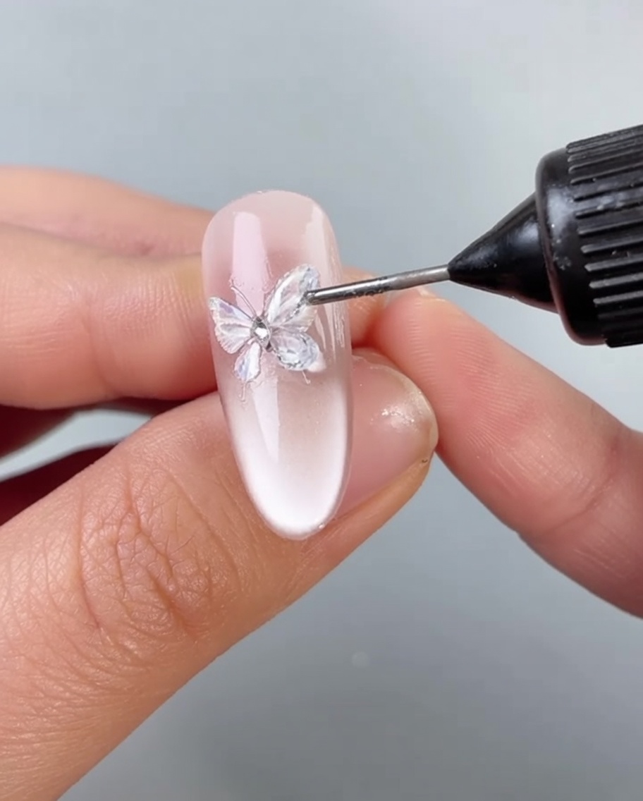 A brush applying a thin layer of clear gel around the base of a butterfly decoration on a nail.