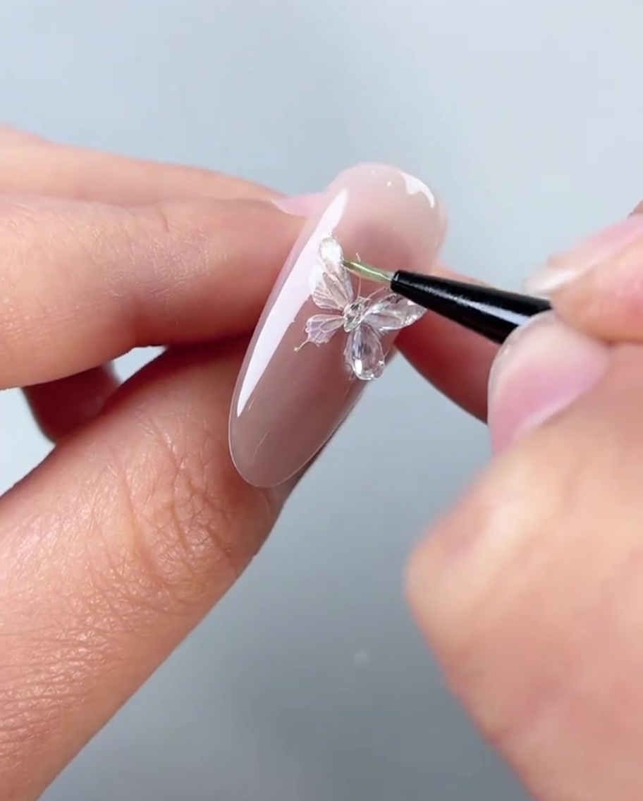 A close-up view of a brush applying thick, clear styling glue onto the left wings of a decorative butterfly sticker on a pink nail.