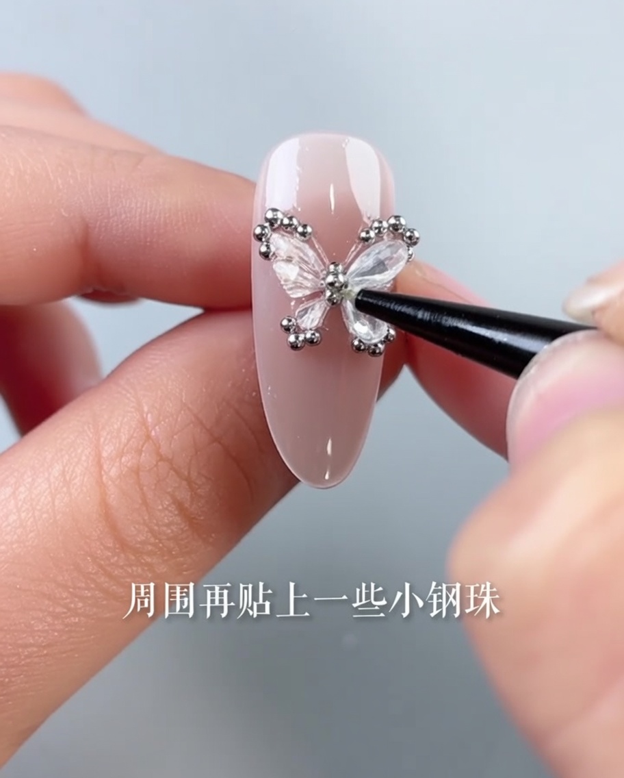 A pair of tweezers placing tiny silver steel beads around the edge of a butterfly-decorated nail.