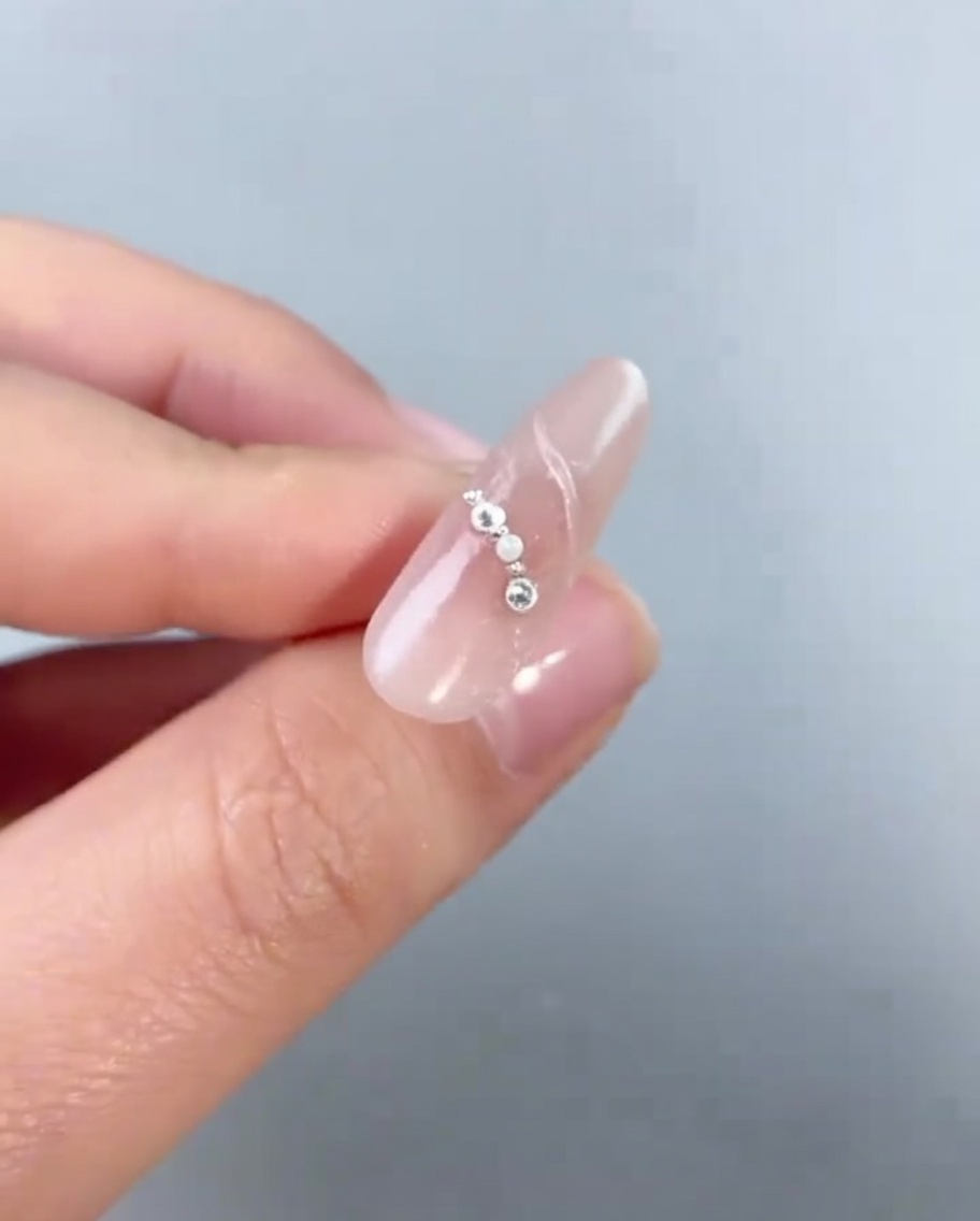 A close-up of a silver pentagram charm being placed onto a nail decorated with small pearls and beads.