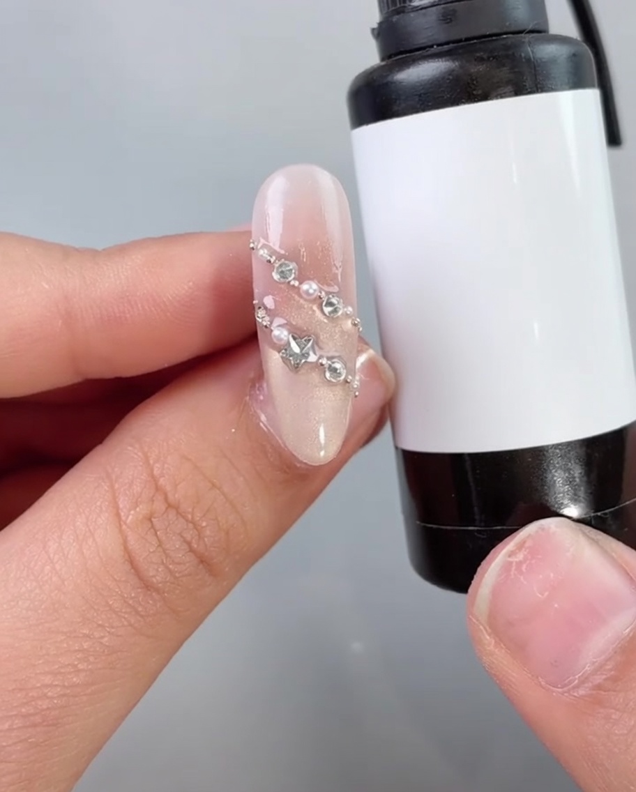 A nail brush applying a clear top coat or sealant over a decorated nail with pearls and diamonds.