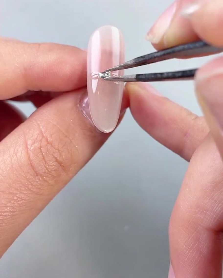 Tweezers placing small silver beads or diamonds horizontally across the middle of a gel-painted nail.