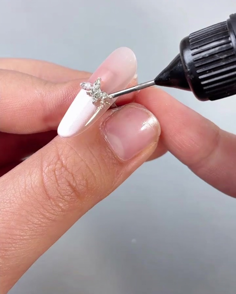 Applying a clear top coat or sealant over a fingernail adorned with a butterfly charm.