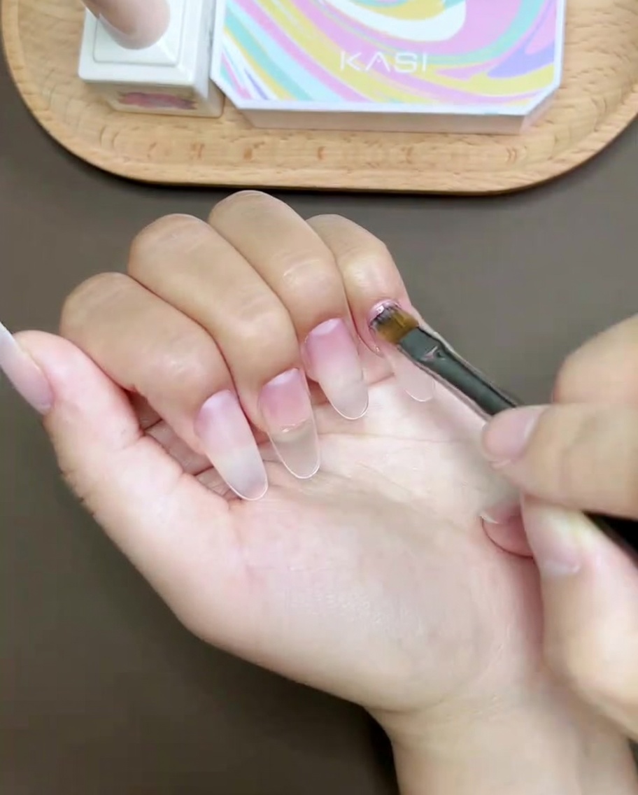 A nail brush painting a pink curved line across the tip of a thumb nail.
