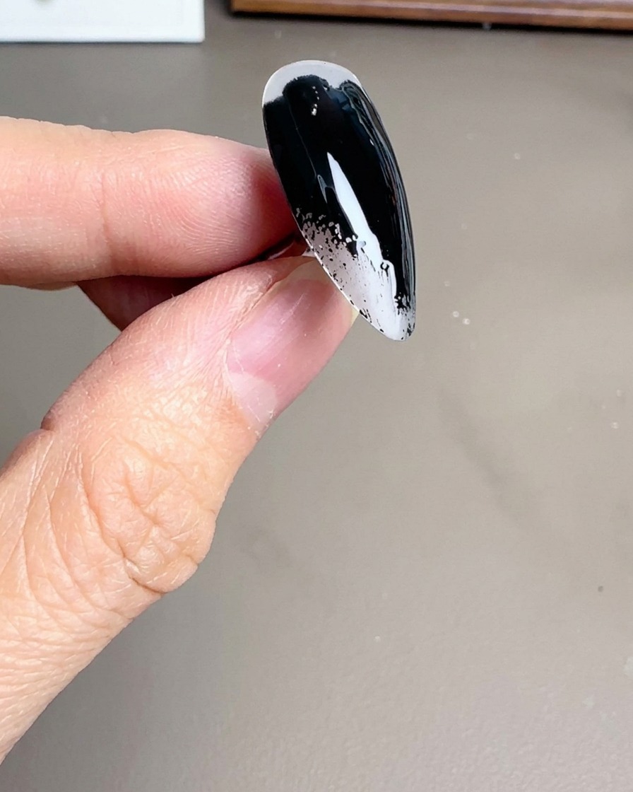 A close-up of a brush applying a glossy black gel polish to a clear stiletto-shaped nail tip.