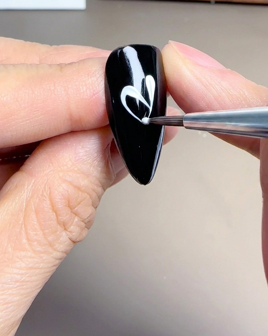A close-up of a fine liner brush creating delicate, elongated white petal shapes on a black nail.