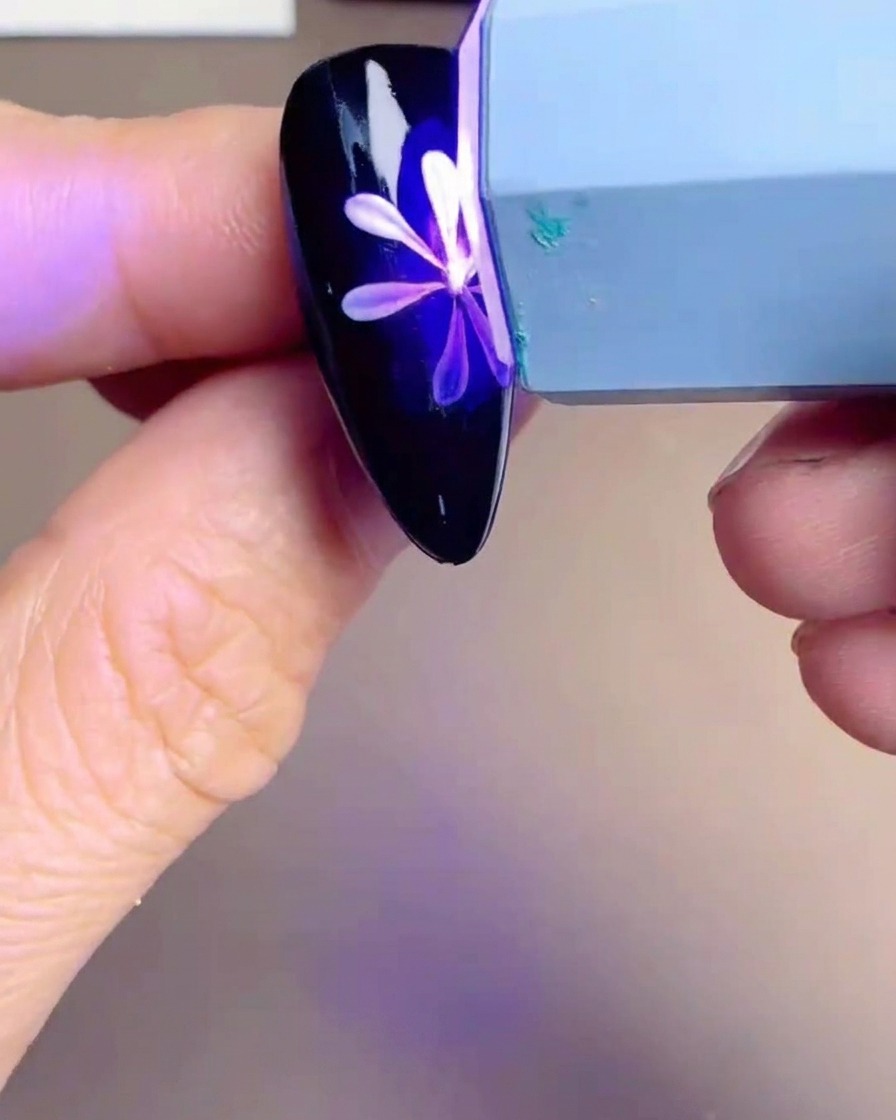 A handheld UV/LED lamp hovering over a nail tip with a white and yellow daisy design, showing a purple glow.
