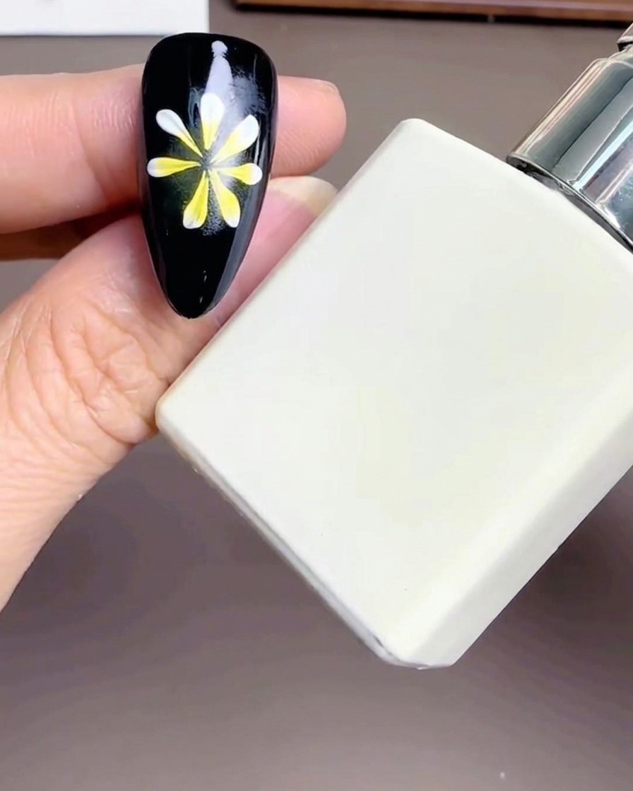 A close-up of a white rectangular bottle of Diamond Top Coat held near a manicured nail with a flower design.
