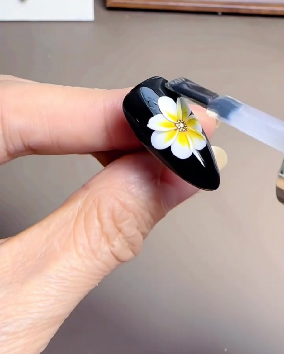 A nail brush applying a clear, glossy gel top coat over a partially finished daisy design on a black nail.