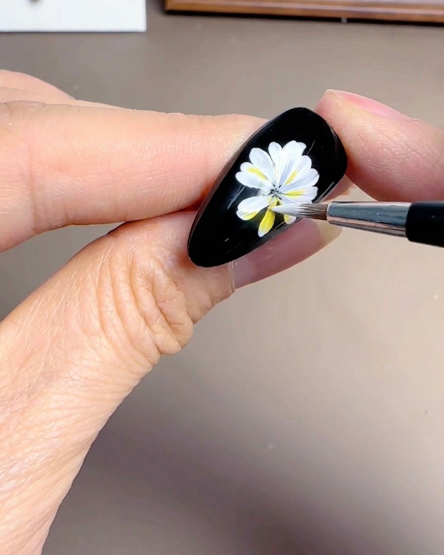 A close-up of a hand adding overlapping white petals to a daisy design on a black nail background.