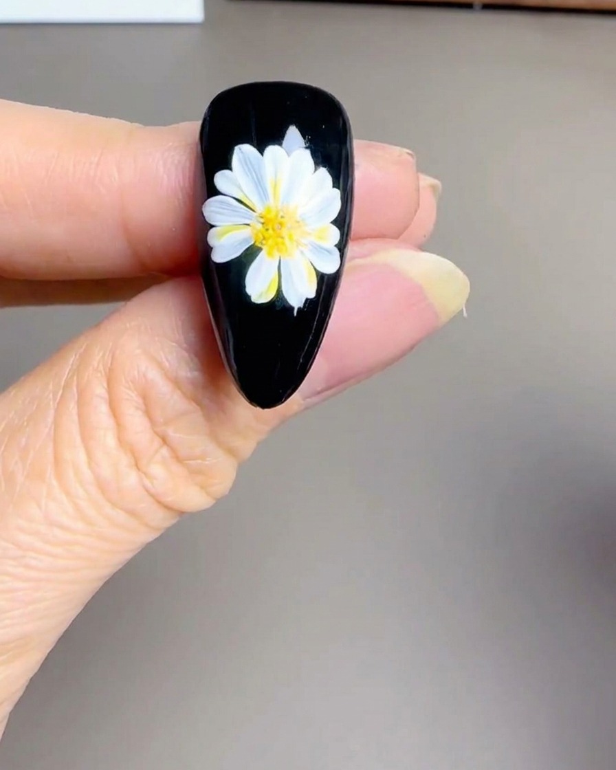 A close-up view of a hand holding a small UV/LED lamp over a finished black nail with a white daisy design.